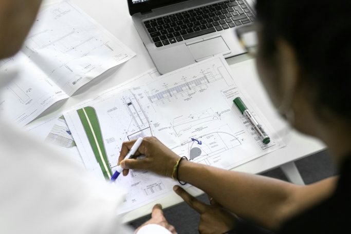 Person schreibt mit einem Stift auf einem Dokument, Laptop im Hintergrund sichtbar.